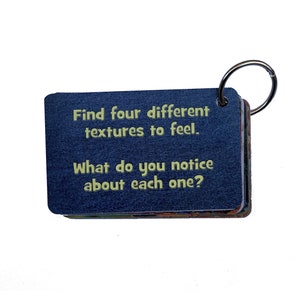 May include: A blue card with a white ring attached to it. The card has the text "Find four different textures to feel. What do you notice about each one?" printed on it.