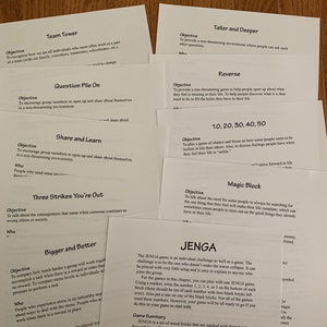 May include: A collection of white papers with black text outlining different team building games. The games include Team Tower, Question Pile On, Share and Learn, Three Strikes You're Out, Bigger and Better, Taller and Deeper, Reverse, 10, 20, 30, 40, 50, Magic Block, and Jenga.