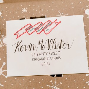 May include: A white envelope with a red and green ribbon design. The envelope is addressed to Kevin McAllister, 23 Fancy Street, Chicago, Illinois 60131. The text "Please Deliver To:" is written on the ribbon.