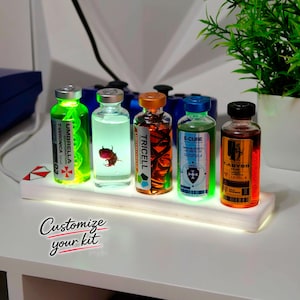 Op de afbeelding: Vijf glazen flesjes met gekleurde vloeistoffen en labels, waaronder Umbrella Corporation T-Veronica, Tricell Uroboros, E-Cure en T-Abyss, geplaatst op een rechthoekige wit verlichte displayvoet. De tekst Customize your kit is zichtbaar.