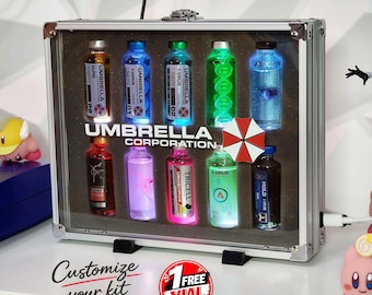 May include: A silver metal briefcase containing ten illuminated glass vials with labels reading Umbrella Corporation, T-Virus, and Tricell. The case features a red and white umbrella logo, a clear front panel, and a power cord extending from the side.