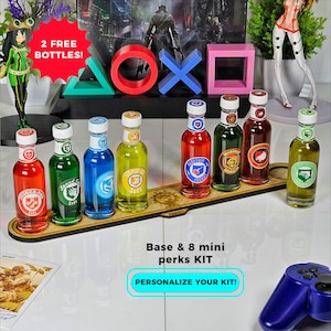 Peut inclure: Un présentoir en bois contenant huit mini bouteilles avec des liquides colorés et des étiquettes. Les bouteilles sont disposées en rangée, avec une variété de couleurs et de motifs. L'image comprend également des symboles PlayStation et une manette.