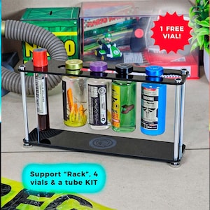 Puede incluir: Un soporte negro que contiene cuatro viales y un kit de tubos. Los viales tienen tapas de colores y contienen varios líquidos. El texto "Support Rack, 4 vials & a tube KIT" está en la parte inferior. Una pegatina roja en forma de estrella dice "1 FREE VIAL!"