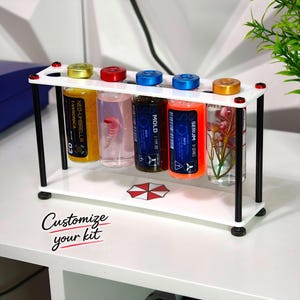 Op de afbeelding: Een wit acryl rek met zwarte steunen met vijf glazen flesjes met gekleurde doppen. De flesjes bevatten diverse vloeistoffen en objecten, met labels zoals Neo-Umbrella T-Veronica, Mold E-Type 001 en Serum U-Series. Een paraplu-logo staat op de basis.
