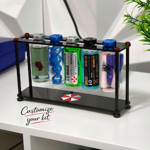 Peut inclure: Un support en acrylique noir contenant cinq flacons en verre avec des liquides colorés. Les flacons portent des étiquettes Umbrella Corporation T-Virus et Neo-Umbrella C-Virus, avec des graphismes d'ADN et d'araignée. La base arbore un logo parapluie.