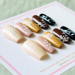 Op de afbeelding: Een set van tien kunstnagels met een paardenbloemdessin. De nagels zijn in verschillende tinten bruin, beige en geel. De nagels zijn in een rij op een wit oppervlak gerangschikt. "We'd love to see your nails!" staat op het oppervlak. "@NailsXLuc&iacute;a" staat ook op het oppervlak.