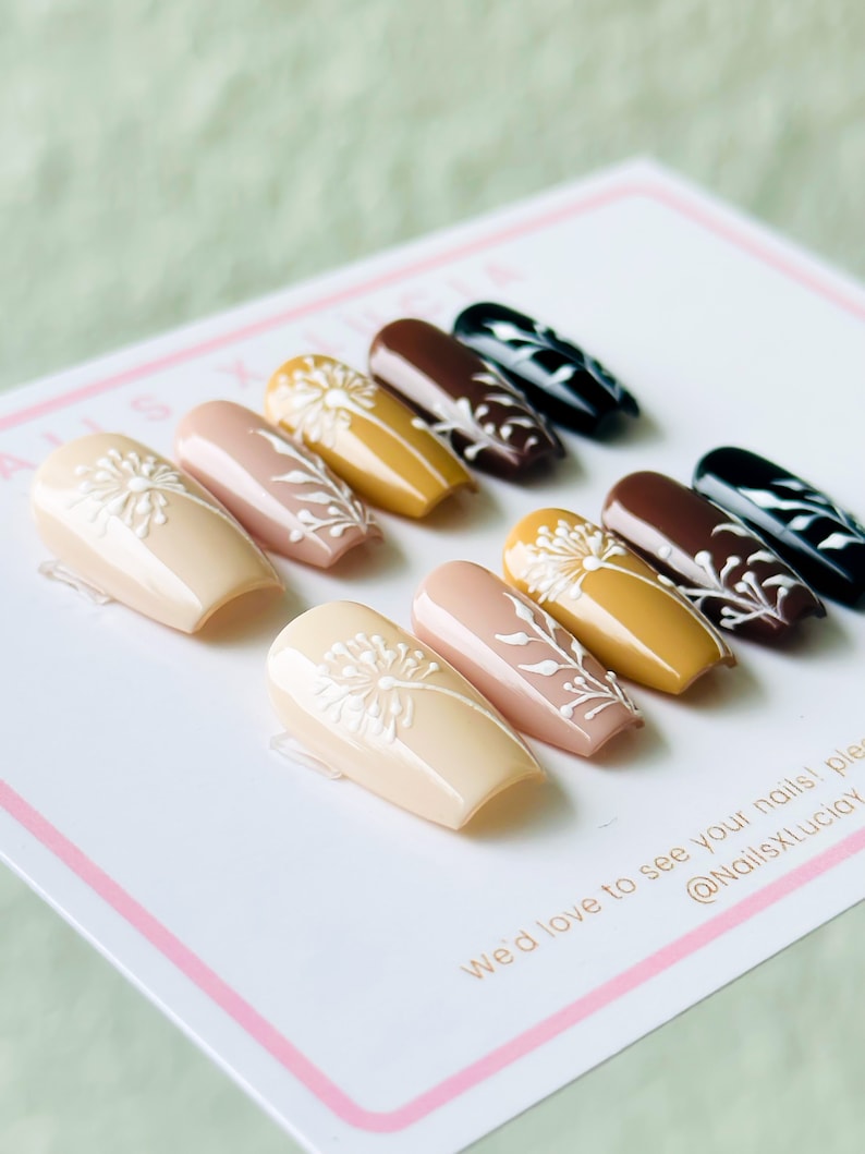 Op de afbeelding: Een set van tien kunstnagels met een paardenbloemdessin. De nagels zijn in verschillende tinten bruin, beige en geel. De nagels zijn in een rij op een wit oppervlak gerangschikt. "We'd love to see your nails!" staat op het oppervlak. "@NailsXLuc&iacute;a" staat ook op het oppervlak.