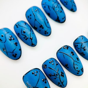Op de afbeelding: Een set blauwe persnagels met een bloemenontwerp. Elke nagel is felblauw geschilderd met zwarte bloemaccenten. De nagels zijn ovaal van vorm en gerangschikt op een wit oppervlak. De tekst "NailsxLucia" is zichtbaar op één nagel.