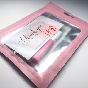 Op de afbeelding: Een roze bedankkaartje met de tekst "Bedankt voor uw bestelling" en een roze-witte sticker met de tekst "Bedankt!" in een roze-witte plastic zak met een transparant venster.