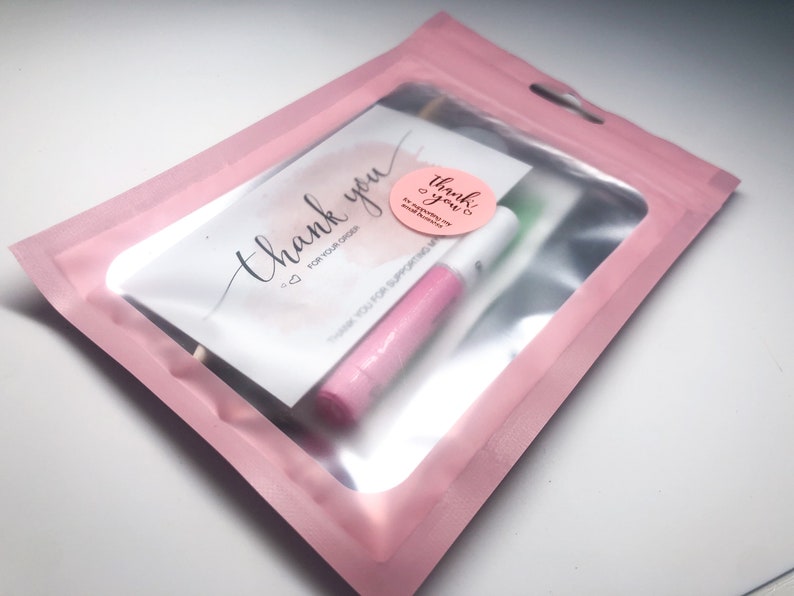 Op de afbeelding: Een roze bedankkaartje met de tekst "Bedankt voor uw bestelling" en een roze-witte sticker met de tekst "Bedankt!" in een roze-witte plastic zak met een transparant venster.