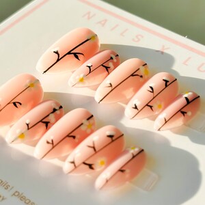Könnte beinhalten: Ein Set mandelförmiger Kunstnägel in zartem Rosa, verziert mit filigranen schwarzen Zweigen und winzigen weißen und gelben Blütenakzenten. Die Nägel sind auf einer weißen Karte mit dem Text "NAILS X LOVE" angeordnet.