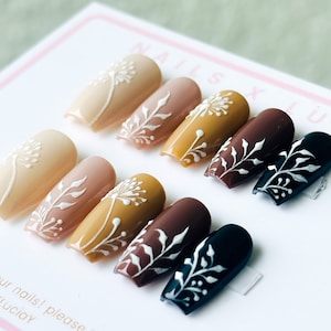 Op de afbeelding: Set van tien kunstnagels met een verloop van nude, bruin en zwart. Elke nagel is versierd met een wit bloemenmotief.