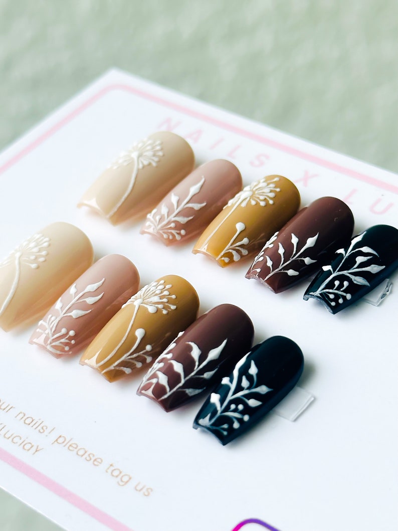 Op de afbeelding: Set van tien kunstnagels met een verloop van nude, bruin en zwart. Elke nagel is versierd met een wit bloemenmotief.