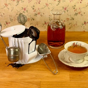 Puede incluir: Un soporte de infusor de té blanco con múltiples infusores de té, una taza de té con un platillo y una jarra de vidrio llena de líquido oscuro. El soporte tiene una tapa y una cadena con un infusor de té redondo colgando. La taza de té es blanca con un patrón floral y está llena de té. La jarra es de vidrio transparente con un fondo redondo y un cuello largo.