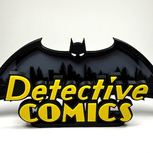May include: A black 3D printed Batman logo with a cityscape silhouette behind it. The words "Detective Comics" are printed in yellow below the logo.