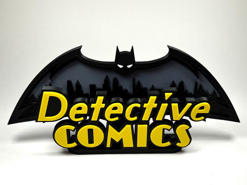 May include: A black 3D printed Batman logo with a cityscape silhouette behind it. The words "Detective Comics" are printed in yellow below the logo.