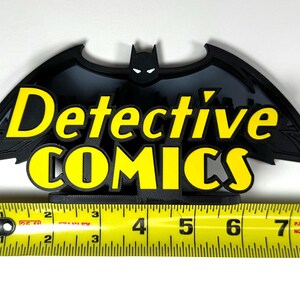 May include: Black 3D printed Batman logo with the words "Detective Comics" in yellow lettering. The logo is approximately 6 inches wide.