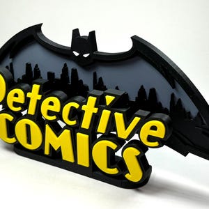 May include: A 3D printed black and yellow sign with the words "Detective Comics" and a silhouette of Batman's bat wings in the background.