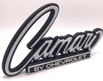 May include: A silver and black plastic emblem with the word "Camaro" in a stylised script font and the words "By Chevrolet" in a smaller font below.