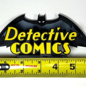 May include: Black 3D printed Batman logo with the words "Detective Comics" in yellow lettering. The logo is approximately 4 inches wide.