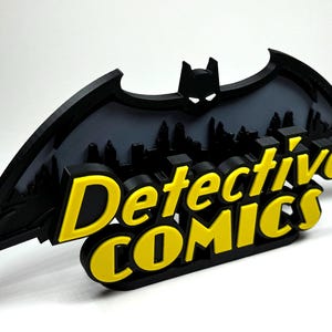 May include: A black 3D printed Batman logo with a cityscape behind it. The words "Detective Comics" are printed in yellow on the logo.