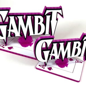 May include: Two 3D-printed signs with the word "GAMBIT" in white, outlined in black, and set against a purple background. The signs are placed on a playing card with the Ace of Spades. The design is a vibrant and eye-catching display.
