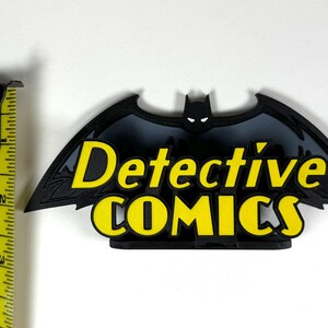 May include: A black and yellow 3D printed logo of a bat with the words "Detective Comics" in yellow letters.