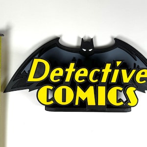 May include: A black and gray 3D printed Batman logo with the words "Detective Comics" in yellow letters.