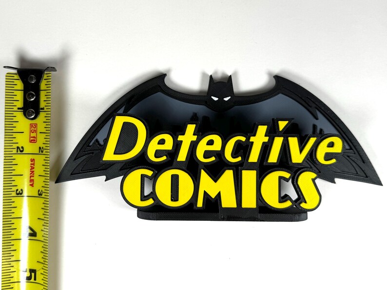 May include: A black and gray 3D printed Batman logo with the words "Detective Comics" in yellow letters.