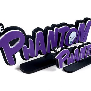 Pode incluir: Logotipo 3D de 'The Phantom' em preto, branco e roxo. O design inclui um gráfico de caveira. O logotipo é montado em uma base preta. O design é uma representação estilizada do personagem de banda desenhada.
