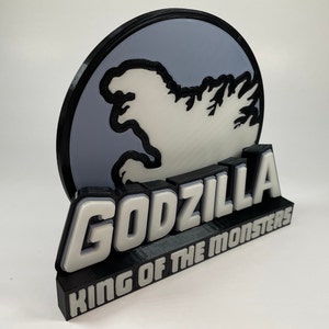 Può includere: Un'insegna di Godzilla stampata in 3D con la scritta "GODZILLA KING OF THE MONSTERS". L'insegna ha una combinazione di colori bianco e nero con uno sfondo grigio e una silhouette di Godzilla.