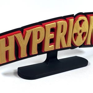 Könnte beinhalten: Ein 3D-gedrucktes Schild mit dem Wort "HYPERION" in goldfarbenen Buchstaben, umrandet in Rot und Schwarz. Das Schild enthält ein stilisiertes Strahlungssymbol. Der Sockel ist schwarz und trägt das Schild.