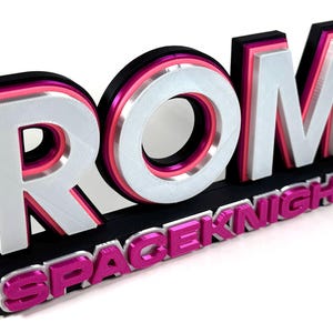 May include: A layered 3D sign featuring the word "ROM" in silver and "SPACEKNIGHT" in pink. The letters are set against a black base with pink and white accents. The design has a modern aesthetic, creating a visually striking effect.
