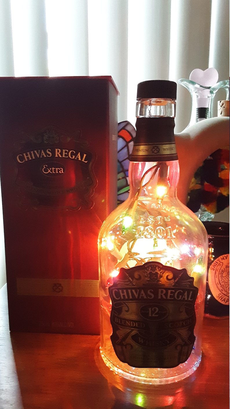 以下が含まれることがあります： 「Chivas Regal 12 Blended Scotch Whisky」と金色に書かれたラベルの付いた透明なガラス瓶が、カラフルな電飾で照らされています。瓶は、「Chivas Regal Extra」と書かれた箱の隣の木製のテーブルの上に置かれています。