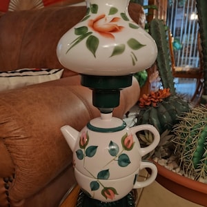 Könnte beinhalten: Eine einzigartige dekorative Lampe mit einem weißen Glasschirm, verziert mit einer handgemalten orangefarbenen Rose. Die Basis ist eine Teekanne und eine Tasse, verziert mit rosa Rosen und grünen Blättern. Der Sockel ist dunkelgrün und quadratisch. Ein charmanter, dekorativer Akzent.