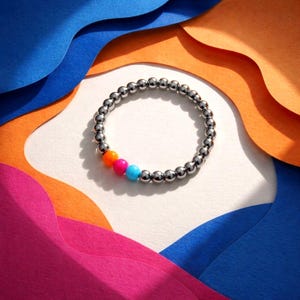 Op de afbeelding: Een zilveren kralenarmband met een gedeelte van oranje, roze en blauwe kralen. De armband ligt op een wit oppervlak met kleurrijke papieren uitsparingen op de achtergrond. De armband is een stretch-stijl.