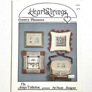 May include: A cross-stitch pattern booklet titled "HeartStrings Country Pleasures" featuring three different designs for pillows and a basket. The designs include a birdhouse, a heart, and a fan.