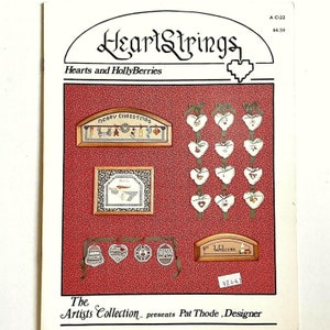May include: A cross stitch pattern booklet titled "HeartStrings: Hearts and HollyBerries" featuring a variety of cross stitch designs including hearts, a welcome sign, and a Christmas sign. The booklet is by The Artists Collection and designed by Pat Thode.