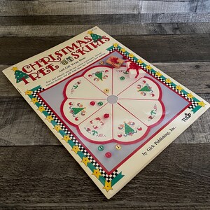 Op de afbeelding: Een boek getiteld "Christmas Tree Skirts and Gift Wrap Ideas" door Gick Publishing, Inc. De omslag toont een witte stoffen boomrok met een rood, groen en wit geborduurd ontwerp. Het boek bevat instructies voor het maken van de boomrok en cadeaupapier.