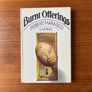 May include: A white book cover with the title "Burnt Offerings" in black text with a decorative border. The author's name, "Robert Marasco", is printed in black text below the title. The book cover features a gold-colored door knob with a human face sculpted into it. The text "A Novel" is printed in black text below the door knob.