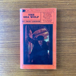 May include: A vintage paperback book cover with an orange background. The title is "The Sea Wolf" by Jack London. The cover features an illustration of a man in a dark coat and hat, looking out from a doorway.
