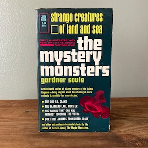 May include: A vintage paperback book cover with a dark blue background and a red title that reads "the mystery monsters". The book is titled "Strange Creatures of Land and Sea" and is by Gardner Soule. The cover features illustrations of sea creatures.