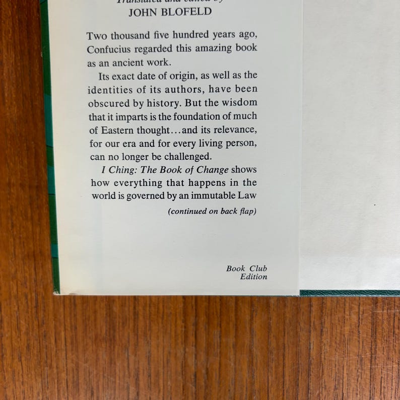 May include: A book cover with text in black font on a white background. The text includes the title "I Ching: The Book of Change" and author John Blofeld. The book is a Book Club Edition. The cover has a green border and is set against a wooden surface.
