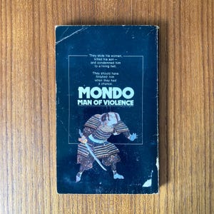 Puede incluir: Portada de libro de bolsillo vintage con el t&iacute;tulo "MONDO MAN OF VIOLENCE". La portada presenta una ilustraci&oacute;n de un guerrero samur&aacute;i con armadura tradicional, sosteniendo una espada. El fondo es azul oscuro con texto blanco.