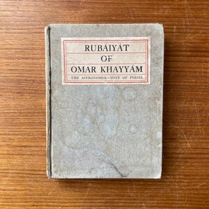 May include: A vintage book titled "Rubáiyát of Omar Khayyám" with a light gray cover and a rectangular label. The label features red borders and black text. The book is on a wooden surface.