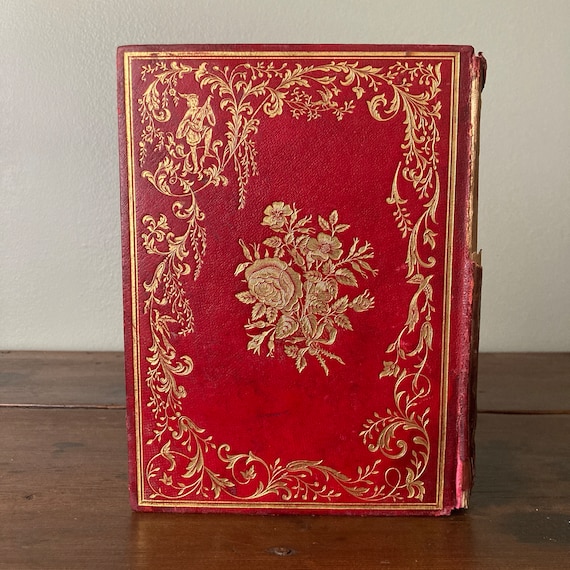 May include: A red leather-bound book with gold-leaf embellishments. The cover features a floral design and a small illustration of a figure playing a musical instrument.
