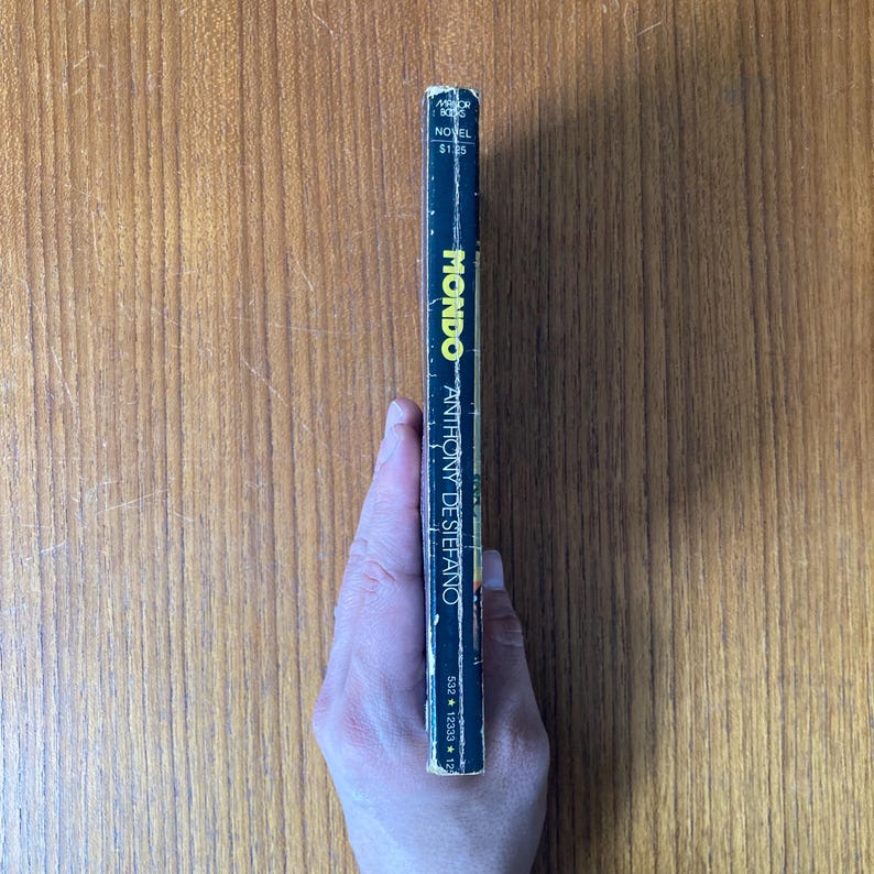 Puede incluir: Un libro de bolsillo vintage titulado "Mondo" de Anthony De Stefano. El lomo es negro con texto amarillo y blanco. El libro est&aacute; sostenido por una mano, con la portada mirando al espectador. El libro est&aacute; sobre una superficie de madera.
