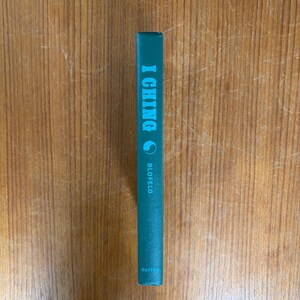 May include: A teal-colored book spine with the title "I Ching" in light blue lettering. The author's name, Blofeld, is printed below the title. The book is set against a wooden background.