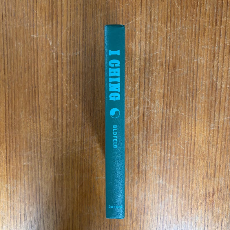 May include: A teal-colored book spine with the title "I Ching" in light blue lettering. The author's name, Blofeld, is printed below the title. The book is set against a wooden background.
