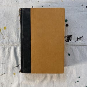 May include: A hardcover book featuring a tan front cover and a black spine. The book has a rectangular shape with a clean, solid-colored surface on the front panel and a vertical black strip along the left side where the binding is located.